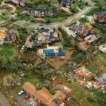 A devastated coastal community in Keaton Beach, Florida, post-Hurricane Helene. The scene shows destroyed homes with debris scattered across the landscape. Some structures are completely flattened, while others are partially standing.