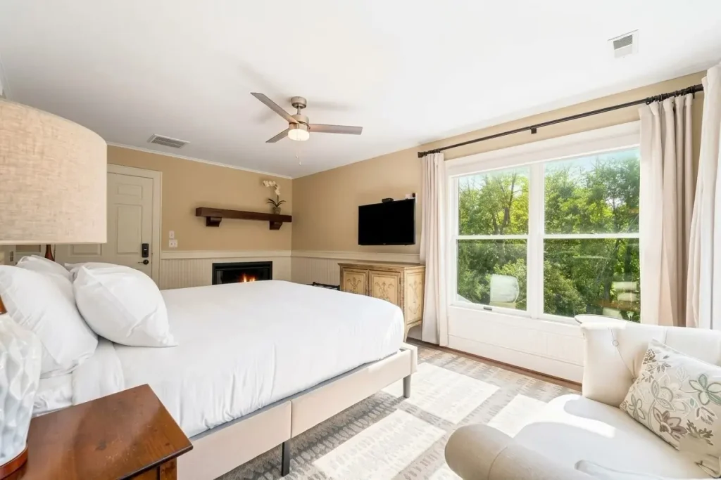 • "Cozy luxury bedroom at the Inn at Brierley Hill featuring a king bed, fireplace, flat-screen TV, cream armchair, and a large window overlooking lush green trees."